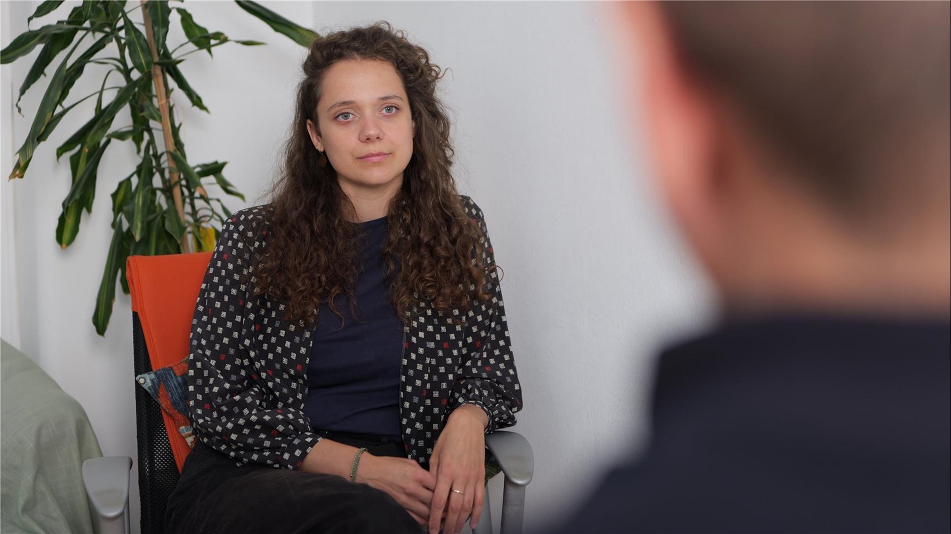 Links im Bild sitzt eine Frau, rechter Bildteil ist die Rückansicht eines Kopfes