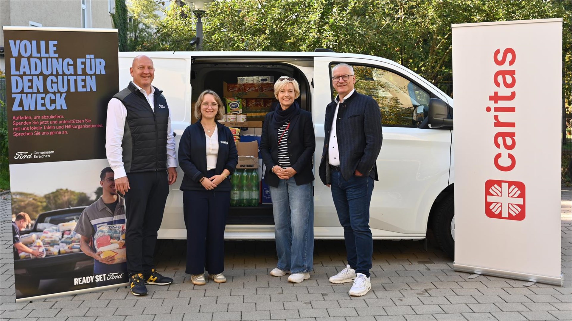 Vier Menschen stehen vor einem weißen Ford Transit. Rechts und links im Bild stehen zwei Roll-Ups