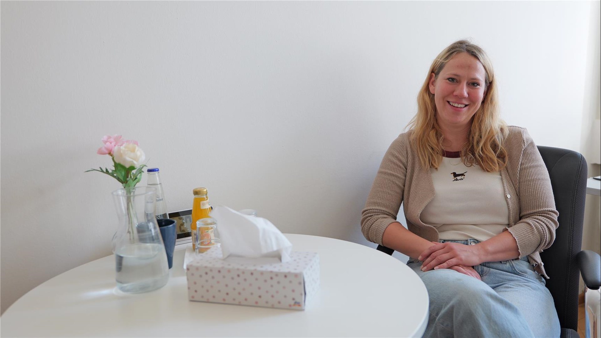 Eine Frau sitzt auf einem Stuhl an einem weißen runden Tisch. Auf dem Tisch steht eine Vase mit zwei Blumen, eine Taschentücherpackung, Getränke und Gläser.